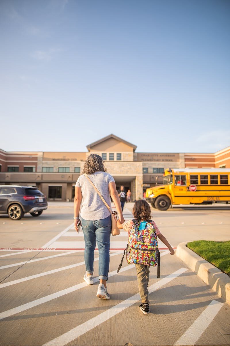 A mother and her daughter walk towards a school building on a sunny day.