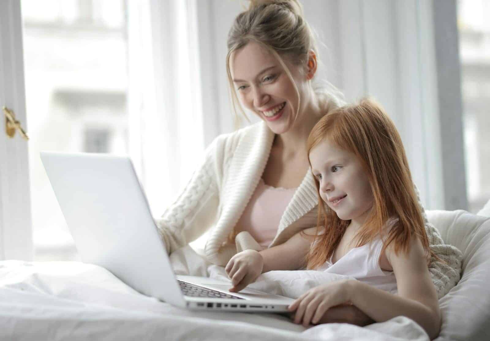 A mother and daughter share a joyful moment watching content on a laptop, basking in the cozy indoor setting.