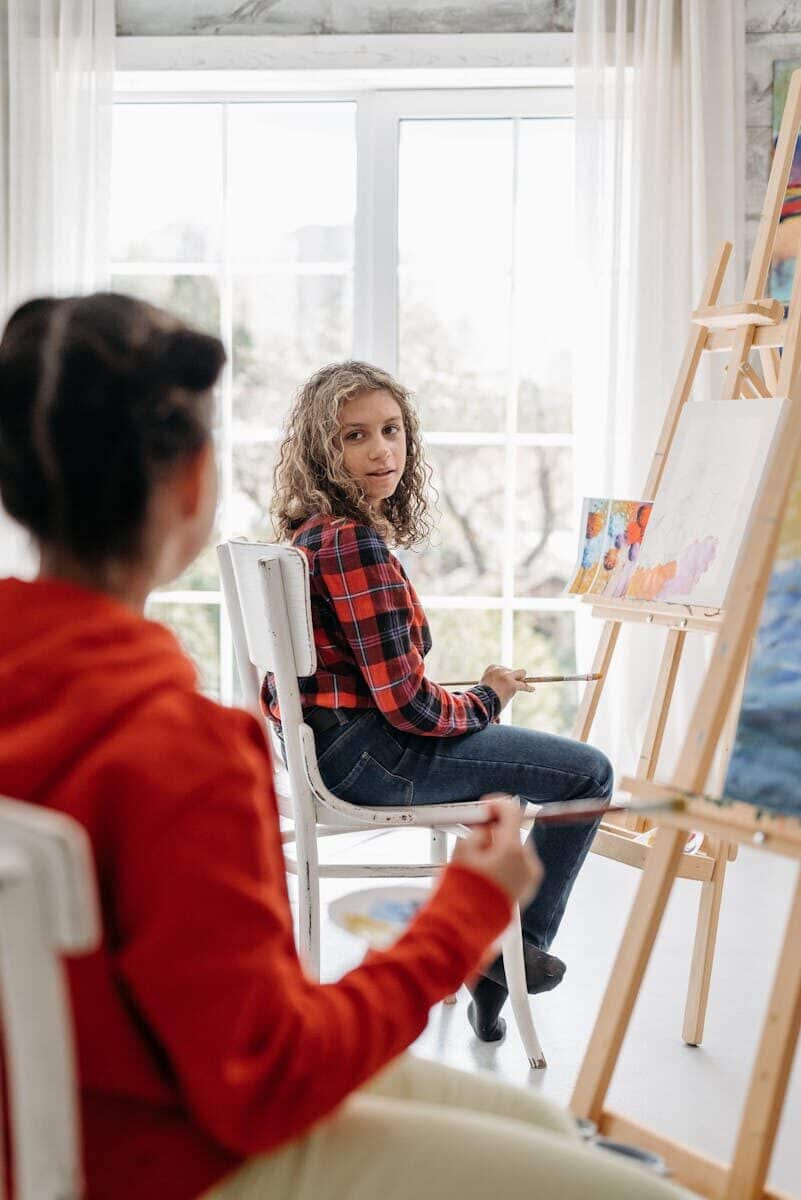 Students in an art class painting at easels, with focus on a woman in a checkered shirt.